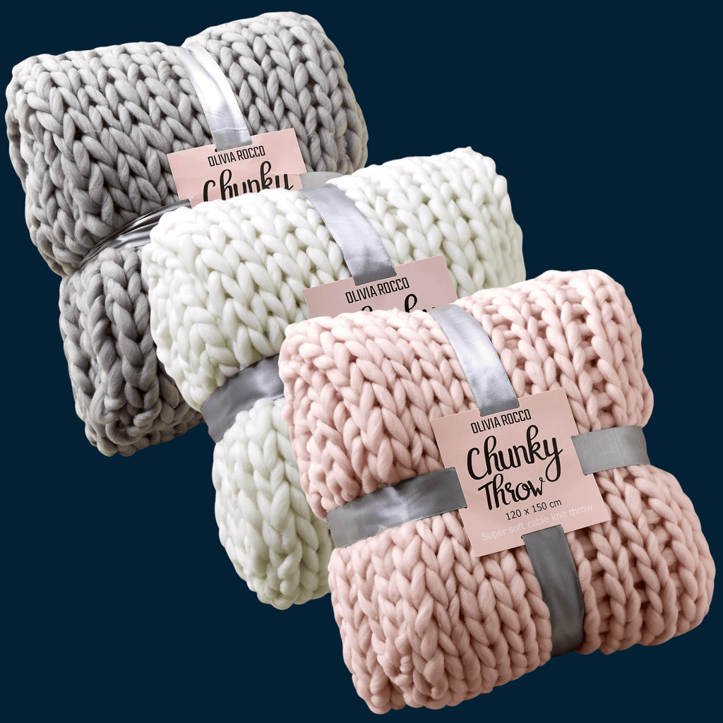Three chunky knit blankets in gray, white, and pink with branded tags on a dark blue background.